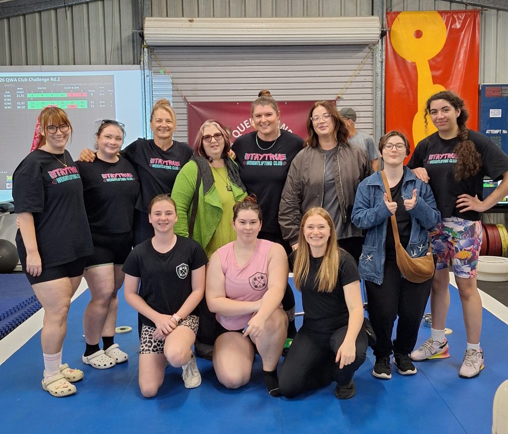 A group of women posing together at a weightlifting club event, wearing matching t-shirts and expressing camaraderie in a gym setting.